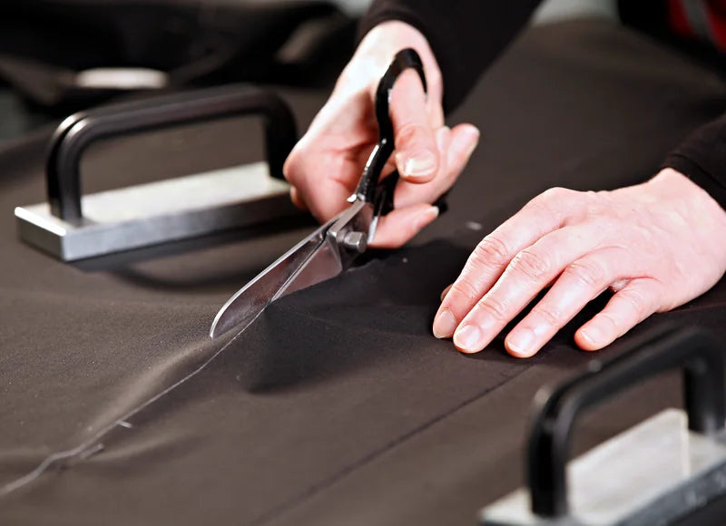 Cutting in the atelier: hands guide the tailor’s shears through black robe cloth, weights hold it on the cutting table